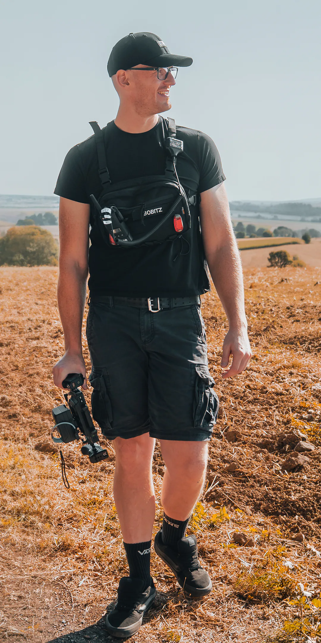 Bastian Obitz mit Kameraausrüstung auf einem abgeernteten Feld, weite Hügellandschaft im Hintergrund