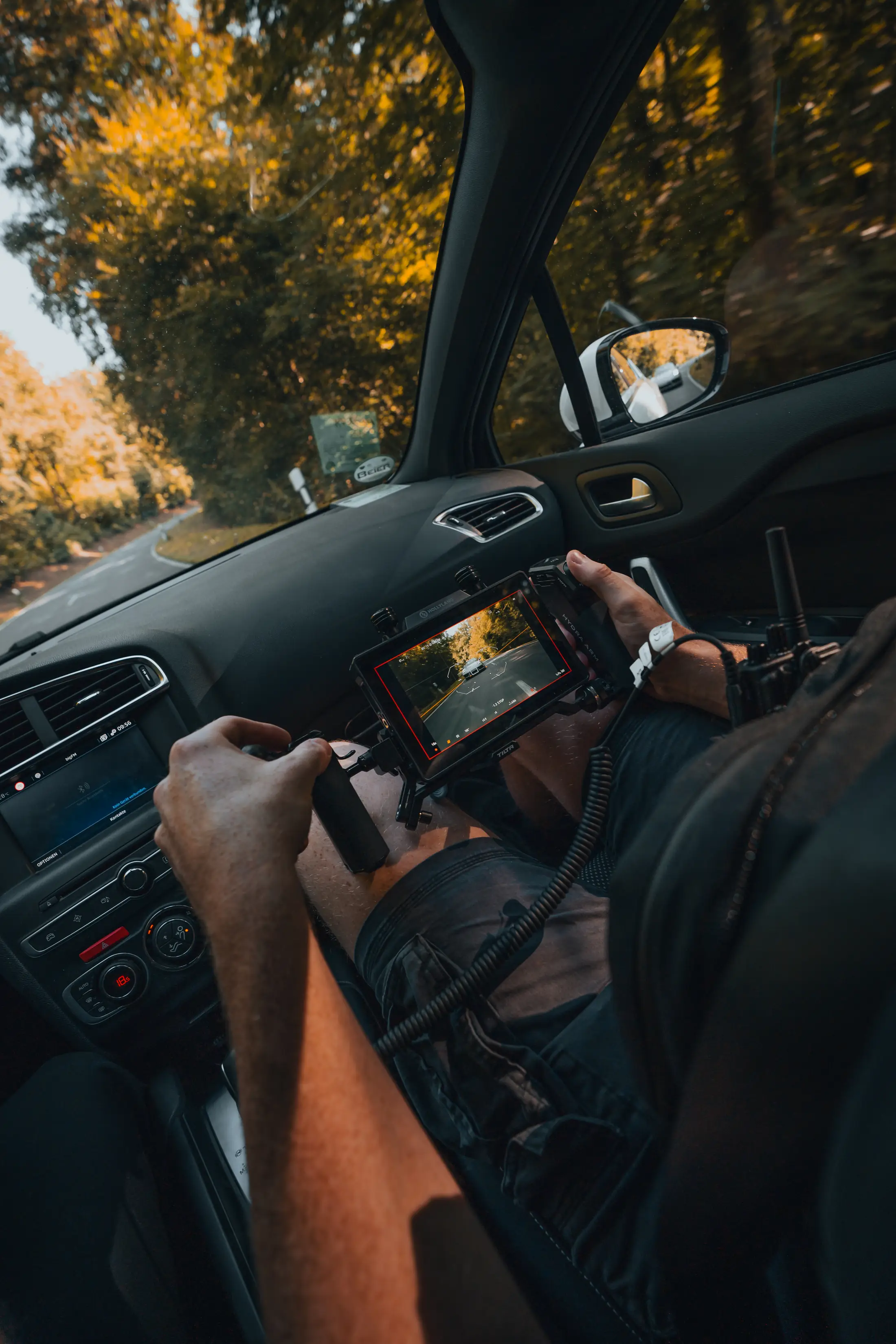 Blick aus dem fahrenden Auto: Hände am Kameramonitor, vorne die Landstraße zwischen herbstlichen Bäumen